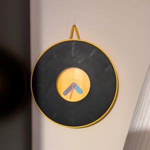 May include: A circular wall clock with a black face and yellow surround. The clock features a yellow centre with red, blue, and yellow hands. It is suspended by a yellow strap.
