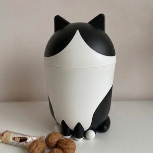 May include: A black and white cat-shaped container with a lid, ears, and paws. The container is cylindrical with a white front and black accents. Walnuts and a candy bar are next to the container.