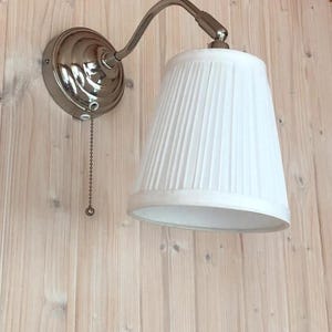 Peut inclure: Une applique murale avec un abat-jour plissé blanc et une finition en nickel brossé. La lampe est équipée d'une chaînette et est fixée sur un mur en bois clair. Le bras de la lampe est courbé.