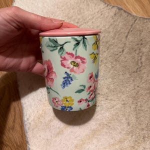 Può includere: Un contenitore cilindrico in ceramica con coperchio rosa. Il contenitore è decorato con un motivo floreale rosa, giallo e blu su sfondo verde chiaro. Il contenitore viene probabilmente utilizzato per la conservazione.