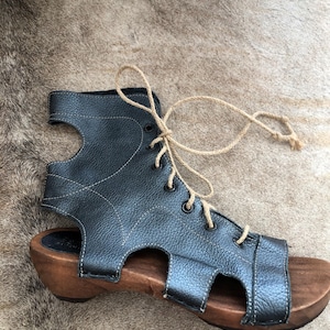 May include: Black leather sandal with a wooden sole. The sandal features a lace-up design with a natural rope-like material. The wooden sole has a slight heel. The sandal has cut-out details on the sides.