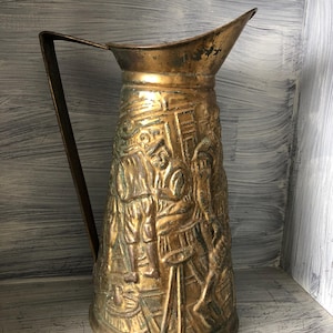 Könnte beinhalten: Eine antike Messingkanne mit einem detaillierten Reliefdesign, das Figuren in historischer Kleidung zeigt. Die Kanne hat einen langen Griff und einen Ausguss. Das Messing hat ein angelaufenes, gealtertes Aussehen, was darauf hindeutet, dass es ein antikes oder Vintage-Objekt ist.