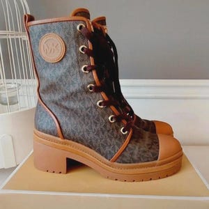 Könnte beinhalten: Ein Schnürstiefel mit dunkelbraunem Monogramm-Print am Schaft, akzentuiert durch hellbraune Lederbesätze und eine abgerundete hellbraune Zehenkappe. Der Stiefel hat eine dicke, profilierte hellbraune Gummisohle, einen Blockabsatz und goldfarbene Ösen.