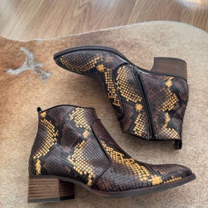 Könnte beinhalten: Ankle Boots mit Schlangenmuster in Braun- und Gelbtönen. Die Stiefel haben einen seitlichen Reißverschluss und einen kleinen Blockabsatz. Die Stiefel werden auf einer hellbraunen Oberfläche präsentiert.