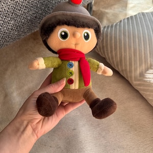 May include: A plush doll wearing a grey hat with a red bobble, brown fur trim, a green jacket, brown trousers, and a red scarf. The doll is being held by a person.