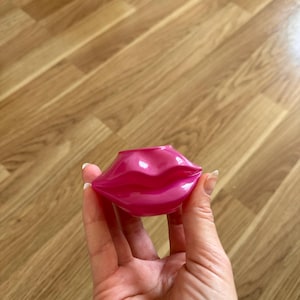 May include: A small, glossy, hot pink container shaped like a pair of puckered lips. The object features a smooth, reflective surface with a defined cupid's bow and full lip contours, designed with a circular opening at the top for storage.