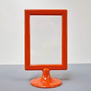 Op de afbeelding: Een helderoranje rechthoekige bordenhouder met een transparant weergavegebied. De bordenhouder heeft een afgeronde basis en een glanzende afwerking. Het frame is feloranje van kleur, geschikt voor het weergeven van borden of advertenties.