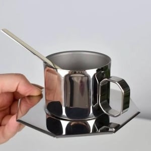 Puede incluir: Una taza de caf&eacute; de acero inoxidable con un platillo y una cuchara a juego. La taza tiene un asa geom&eacute;trica. El juego es muy reflectante, con un acabado pulido. Una mano sostiene el juego.