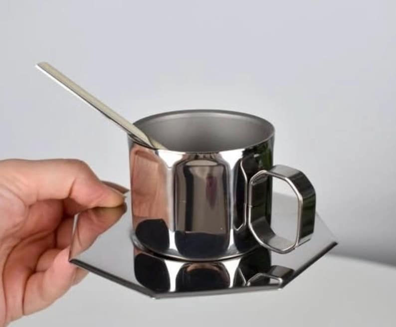 Puede incluir: Una taza de caf&eacute; de acero inoxidable con un platillo y una cuchara a juego. La taza tiene un asa geom&eacute;trica. El juego es muy reflectante, con un acabado pulido. Una mano sostiene el juego.