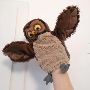 May include: A brown owl hand puppet with outstretched wings. The puppet has large yellow eyes, a tan chest, and brown wings and head. The puppet's feet are gray. The puppet is made of soft, plush material.
