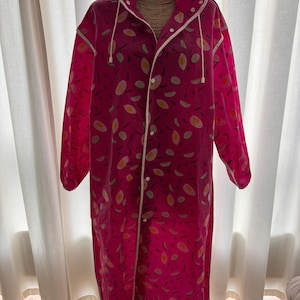 Può includere: Un impermeabile rosa acceso con cappuccio, bordi bianchi e chiusura con bottoni. Il cappotto presenta un motivo ripetuto di disegni a forma di uovo in giallo, bianco e grigio. L'impermeabile è progettato per essere indossato sopra i vestiti.