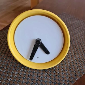 May include: A yellow-rimmed clock with a white face and black hands. The clock's hands are positioned to indicate the time. The clock is a simple, modern design, suitable for home or office decor.