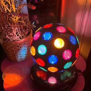 May include: A black disco ball lamp with colorful, circular lights in red, blue, yellow, pink, and green. The lamp sits on a black base and a pink, flower-shaped stand. A wicker basket with plants is to the left.