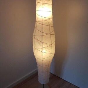 May include: A tall floor lamp featuring a cylindrical, slightly tapered white paper shade wrapped in thin, crisscrossing cord detailing. The lamp emits a warm, diffused glow and is supported by a circular, metallic base on a thin vertical stand.