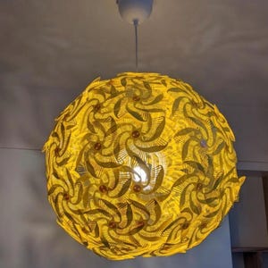 Könnte beinhalten: Eine kugelförmige, gelbe Pendelleuchte mit einem komplexen, geschichteten Design. Das Licht wird von einer weißen Deckenleuchte abgehängt. Das komplizierte Muster erzeugt ein warmes, einladendes Leuchten, ideal für die Inneneinrichtung.