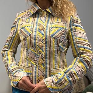May include: A colourful jacket with a collared neckline and large, round buttons. The jacket features a mix of vertical stripes and plaid patches in yellow, blue, and brown. The jacket has two front pockets and decorative stitching.