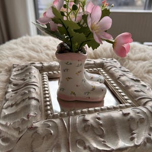 Peut inclure: Une petite botte en céramique blanche avec des accents roses et un motif floral est remplie d'un bouquet de fleurs roses. La botte est posée sur un cadre orné blanc avec un miroir.