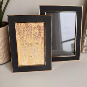 May include: Two black picture frames with gold trim. The smaller frame has a gold background with the text "Rachel Zoe" and "Los Angeles, New York" printed on it. The larger frame is empty.