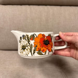 Puede incluir: Una salsera de cerámica blanca con un diseño floral con amapolas naranjas, margaritas blancas y hojas verdes.