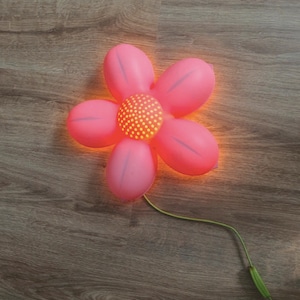 Puede incluir: Una luz nocturna de color rosa en forma de flor con un centro amarillo y un cable verde.