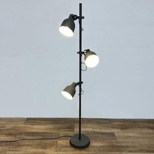 Peut inclure: Un lampadaire gris foncé avec trois spots réglables. La lampe a une base ronde et un poteau vertical avec les spots fixés à différentes hauteurs. Les spots sont orientés vers le bas et la lampe est branchée sur une prise électrique.