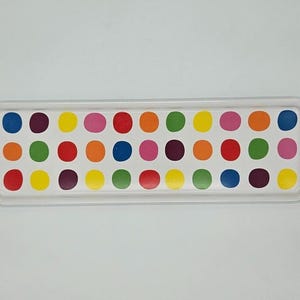 Puede incluir: Una bandeja rectangular blanca con un colorido estampado de lunares. Los puntos están dispuestos en filas y columnas, con una variedad de colores que incluyen rojo, amarillo, verde, azul, naranja y rosa. La bandeja tiene un borde elevado.