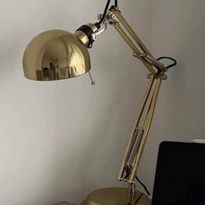 May include: A gold-toned metal desk lamp with a polished dome-shaped shade, an adjustable articulated arm, and a circular base. The lamp features black accents at the joints and a black power cord, providing a functional lighting solution.