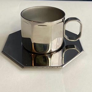 Puede incluir: Un juego de taza y platillo de caf&eacute; de acero inoxidable. La taza tiene un dise&ntilde;o geom&eacute;trico con un asa a juego. El platillo es de forma hexagonal, de color gris oscuro. El conjunto es reflectante.