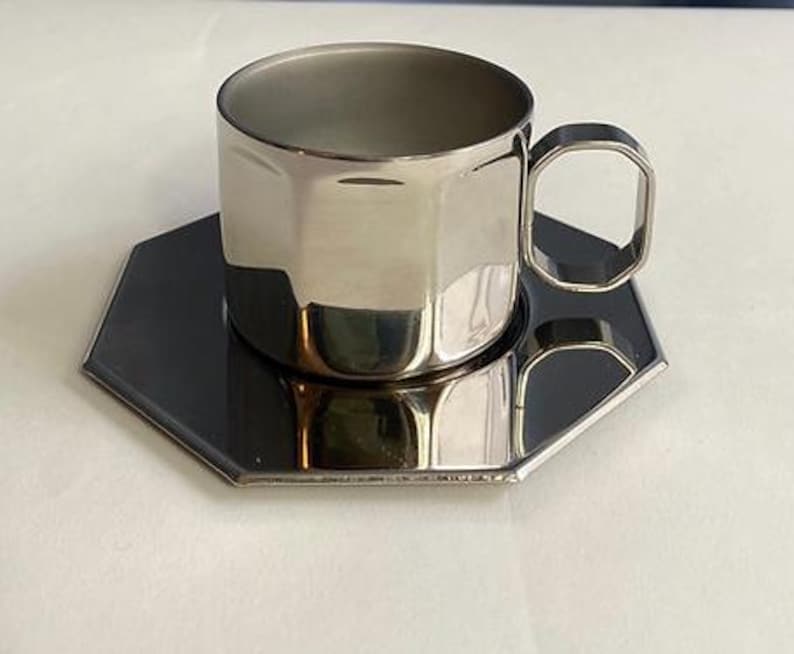 Puede incluir: Un juego de taza y platillo de caf&eacute; de acero inoxidable. La taza tiene un dise&ntilde;o geom&eacute;trico con un asa a juego. El platillo es de forma hexagonal, de color gris oscuro. El conjunto es reflectante.