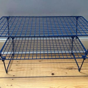 May include: A two-tier stackable wire shelf unit featuring a vibrant blue finish. The structure is composed of a rectangular grid pattern made from thin metal wire, supported by four foldable legs on each level, creating an open storage design.