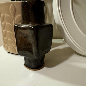 Può includere: Un vaso in ceramica marrone scuro con corpo quadrato e base arrotondata. Il vaso ha una superficie testurizzata e una finitura leggermente irregolare.