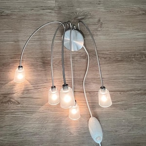 May include: A modern ceiling light fixture with six frosted glass shades. The fixture has a brushed metal finish and curved arms. The light is turned on, illuminating the shades. The light has a white cord and a white on/off switch.