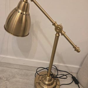 May include: A brass-colored desk lamp with an adjustable arm and a conical shade. The lamp has a round base and a black power cord. The shade has small ventilation holes. The lamp is designed for task lighting.