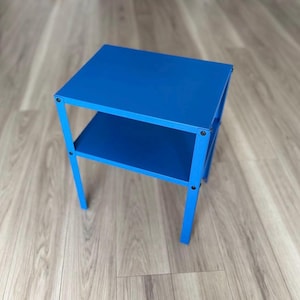 Puede incluir: Una mesa auxiliar azul brillante de dos niveles. La mesa tiene una parte superior cuadrada y un estante inferior, sostenidos por cuatro patas rectas. La mesa es de metal y está pintada de color azul sólido.