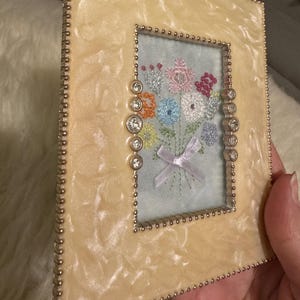 May include: A rectangular picture frame with a cream-colored, marbled finish and a beaded border. The frame holds a small embroidered bouquet of colorful flowers on a light blue background. Clear, round jewels are set on the sides.
