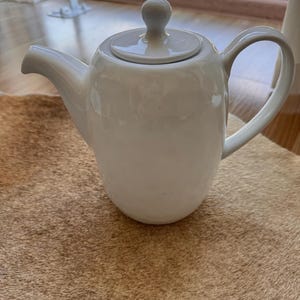 以下が含まれることがあります： カーブしたハンドル、注ぎ口、蓋が付いた白いセラミックティーポット。 ティーポットは光沢があり、茶色のテクスチャのある表面に置かれています。 デザインはシンプルでエレガントで、お茶を出すのに適しています。