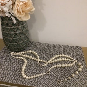 May include: A set of three pearl necklaces on a patterned gray surface. The necklaces are made of white pearls and are of varying lengths.