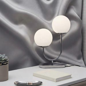 May include: A table lamp featuring two spherical white glass shades supported by a polished chrome U-shaped metal frame and an oval base. The lamp stands on a white surface next to a closed notebook, a small succulent plant, and a metal tray.