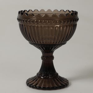 Può includere: Una ciotola in vetro marrone scuro con bordo smerlato e dettagli a coste verticali. La ciotola ha una base rotonda e uno stelo decorativo. Il vetro è traslucido.
