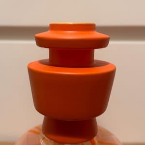 Puede incluir: Primer plano de un tapón de botella naranja. El tapón tiene forma cilíndrica con una base acampanada y una parte superior más pequeña y escalonada. El color es naranja vibrante y sólido, y el diseño es moderno y minimalista.
