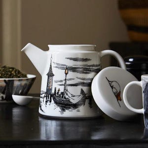 May include: A white ceramic teapot featuring a black and white ink-style illustration of a boat on water near a tall structure. The lid, placed beside the teapot, displays a small orange and black graphic of a creature with a long snout.