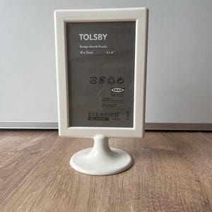 May include: White picture frame with a stand, designed by Henrik Preutz. The frame is 10 x 15 cm or 4 x 6 inches. The frame has a label that says "TOLSBY" and "IKEA".