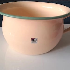 Puede incluir: Un orinal de esmalte de color melocotón con borde y asa verdes. El orinal tiene forma redondeada y un pequeño logotipo en el lateral. El orinal está sobre una superficie blanca.