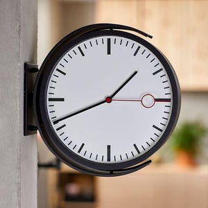 Op de afbeelding: Een dubbelzijdige wandklok met een zwarte ronde omlijsting en een witte wijzerplaat. De klok heeft zwarte uur- en minuutmarkeringen, zwarte wijzers en een dunne rode secondewijzer met een cirkelvormig rood detail nabij de punt van de wijzer.