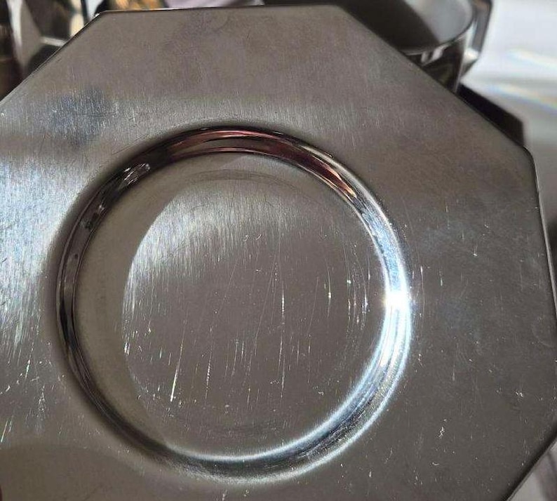 Puede incluir: Primer plano de un plato octogonal plateado con una hendidura circular en el centro. El plato tiene una superficie brillante y reflectante con ara&ntilde;azos visibles.