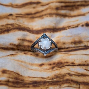 Op de afbeelding: Een zilveren ring met een facetgeslepen, ronde, witte edelsteen. De ring heeft een unieke, getextureerde band en een decoratieve zetting. De ring is geplaatst tegen een achtergrond van een bruin en tan patroon.
