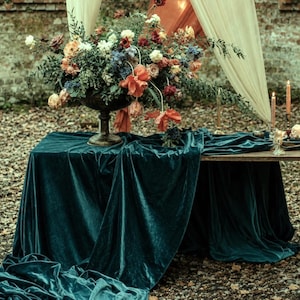 Puede incluir: Una mesa rectangular cubierta con un mantel de terciopelo azul cerceta profundo, con un gran arreglo floral en un jarrón de pedestal oscuro, varias velas encendidas en candelabros dorados y cortinas de tela crema y naranja al fondo.
