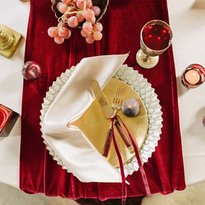 Pode incluir: Um prato de serviço dourado com uma borda branca está sobre um corredor de mesa de veludo vermelho. Um guardanapo branco está dobrado sobre o prato, com uma faca e um garfo dourados colocados em cima. Um único figo está no prato.