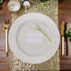 Op de afbeelding: Een wit bord met een gouden rand en een witte bloem op een gouden glitter tafelkleed. Het bord heeft een plaatskaart met de tekst "Bride".