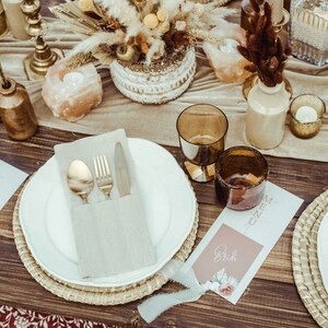 Op de afbeelding: Een rustieke tafelsetting met een wit bord, gouden bestek en een beige servet. De tafel is gedecoreerd met bruine rieten placemats, bruin en gouden glaswerk en een bloemstuk. De plaatskaarten zijn bedrukt met de namen "Bride" en "Groom".